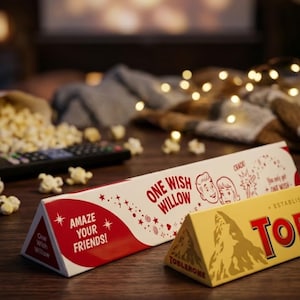 Puede incluir: Una caja triangular blanca etiquetada como One Wish Willow con texto rojo e ilustraciones de dos rostros, colocada junto a una caja de chocolate Toblerone triangular amarilla. El empaque presenta las frases Amaze Your Friends y Crack.