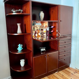 Könnte beinhalten: Ein großes modulares Wandmöbel aus dunklem Holz mit einem geschwungenen offenen Regalbereich auf der linken Seite, einem zentralen Barschrank mit klappbarer Spiegelfläche, Glasflaschen und Stielgläsern sowie einem rechten Teil mit Schrank und fünf Schubladen.