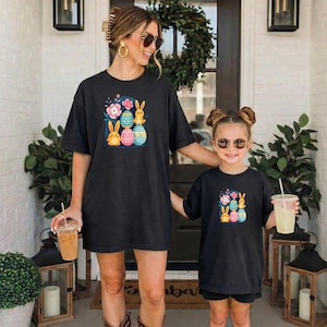 May include: Black t-shirts with Easter-themed graphics. The shirts feature colorful illustrations of Easter eggs, bunnies, and flowers. The adult shirt is oversized, and the child's shirt is a smaller version of the same design.