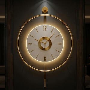 May include: A modern wall clock with a gold-colored frame and accents. The clock face is white with gold hands and numerals. The clock has a circular design with a pendulum and is mounted on a dark gray wall.