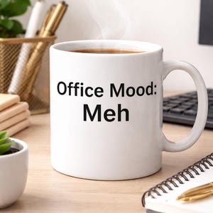 May include: White ceramic mug with the text "Office Mood: Meh" in black. The mug is filled with a dark liquid, possibly coffee or tea, and is on a wooden desk. A spiral notebook and pen are nearby.