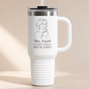 May include: A white stainless steel tumbler with a handle, a lid, and a straw. The front features a black line drawing of a person holding a book, with the text Mrs. Smith, TEACHING THROUGH HOT FLASHES printed in black lettering below the illustration.