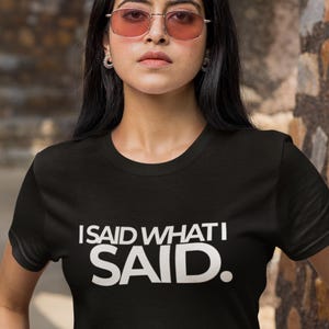 Könnte beinhalten: Schwarzes T-Shirt mit Rundhalsausschnitt und dem weißen Text "I SAID WHAT I SAID." Das Shirt hat eine schwarze Farbe und der Text ist in einer fetten, serifenlosen Schriftart gehalten. Das Model trägt rosafarbene Sonnenbrillen.
