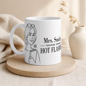 May include: A white ceramic mug featuring a black line art illustration of a person with glasses holding a book. The text printed on the mug reads: Mrs. Smith TEACHING THROUGH HOT FLASHES. The mug has a curved handle and a smooth, glossy finish.
