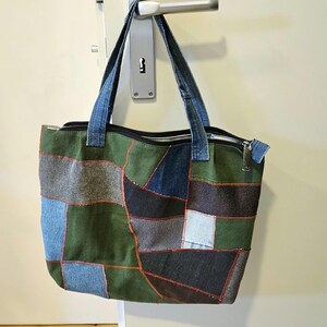 Könnte beinhalten: Eine Patchwork-Tragetasche mit Reißverschluss und Jeans-Henkeln. Die Tasche besteht aus verschiedenen Jeans- und grünen Stoffflicken, die mit rotem Faden zusammengenäht sind. Die Tasche hängt an einem Türgriff.