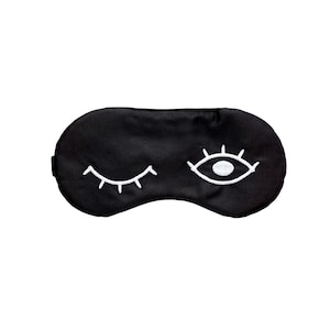May include: A black sleep mask with white embroidered eye designs. One eye is open, and the other is closed, with a curved eyelash design. The mask is made of a soft, dark fabric, perfect for blocking out light.