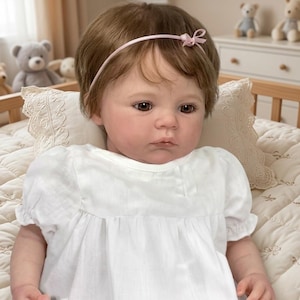 Puede incluir: Una muñeca realista con ojos marrones y cabello castaño corto, vestida con un vestido blanco y una diadema rosa. La muñeca está acostada sobre una manta acolchada con una almohada decorativa. Se ven juguetes blandos en el fondo.