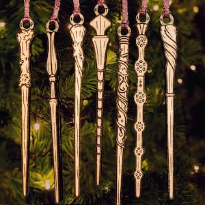 May include: Six gold-toned metal wand ornaments hanging from thin red ribbons. Each wand features a distinct, intricate design with unique textures, carvings, and shapes, including spiral, knobby, and smooth patterns, suspended vertically in a row.