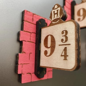 May include: A wooden magnet featuring a pink brick wall section and a sign displaying the number 9 3/4 with a small H crest at the top. The sign is attached to a black vertical mount, and the text is engraved in a dark brown colour on light wood.