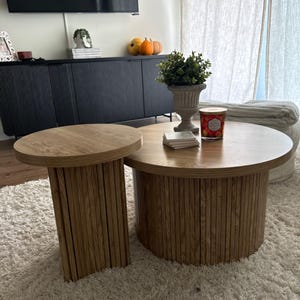 May include: Two wooden coffee tables with a light brown finish. The larger table is round, and the smaller table is rectangular. Both tables have a slatted design on the sides. The tables are on a white rug.