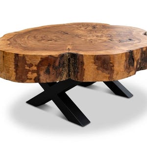 Peut inclure: Une table basse ronde en bois naturel avec un bord brut. Le plateau est de couleur brun clair avec un grain de bois visible. Elle est soutenue par une base en métal noir en forme de X. La table mesure environ 91 cm de diamètre.