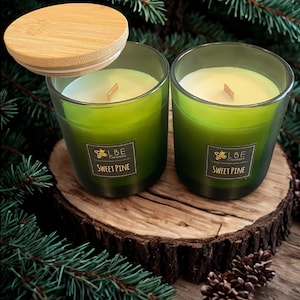 May include: Two green-tinted glass candles with wooden wicks and lids. The candles are labeled "Sweet Pine" and sit on a wooden surface, surrounded by pine needles and cones. The lid of one candle is slightly ajar.
