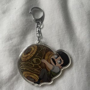 May include: A clear acrylic keychain featuring a cartoon character with a mustache hugging a brown and gold orb. The keychain has a silver clasp and chain.