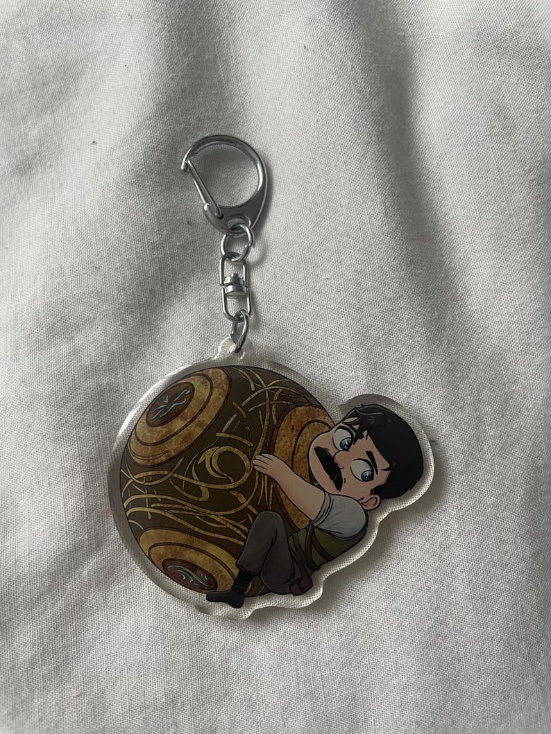May include: A clear acrylic keychain featuring a cartoon character with a mustache hugging a brown and gold orb. The keychain has a silver clasp and chain.