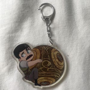 May include: A clear acrylic keychain featuring a cartoon character with a mustache hugging a brown, circular object with a detailed design. The keychain has a silver-colored clasp and chain.
