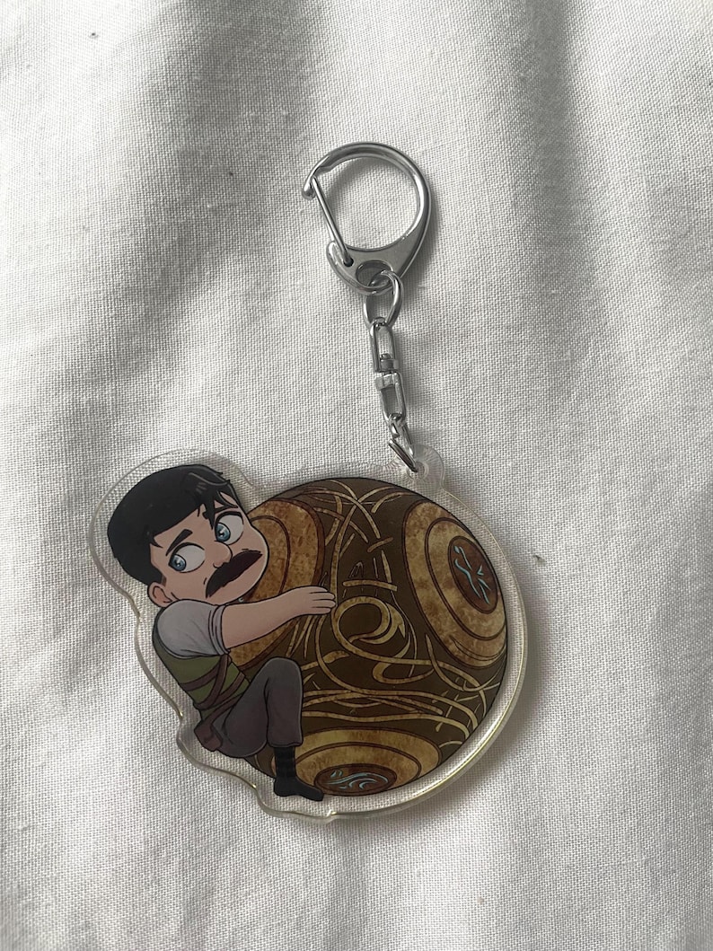 May include: A clear acrylic keychain featuring a cartoon character with a mustache hugging a brown, circular object with a detailed design. The keychain has a silver-colored clasp and chain.