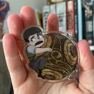 May include: A clear acrylic keychain featuring a cartoon illustration of a man with a mustache clinging to a brown and gold orb. The man is wearing a green vest, gray pants, and a white shirt. The orb has intricate swirling patterns.