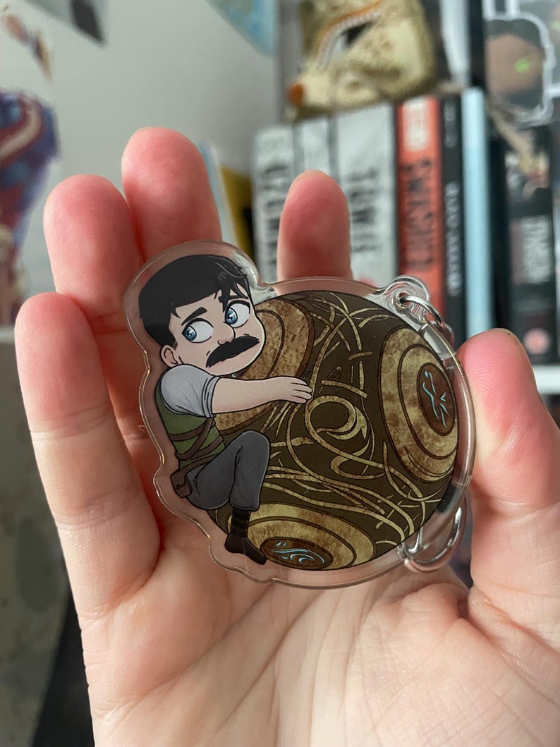 May include: A clear acrylic keychain featuring a cartoon illustration of a man with a mustache clinging to a brown and gold orb. The man is wearing a green vest, gray pants, and a white shirt. The orb has intricate swirling patterns.
