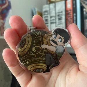 May include: A clear acrylic keychain featuring a cartoon illustration of a character with a mustache hugging a golden, patterned sphere. The keychain has a silver metal ring for attachment.