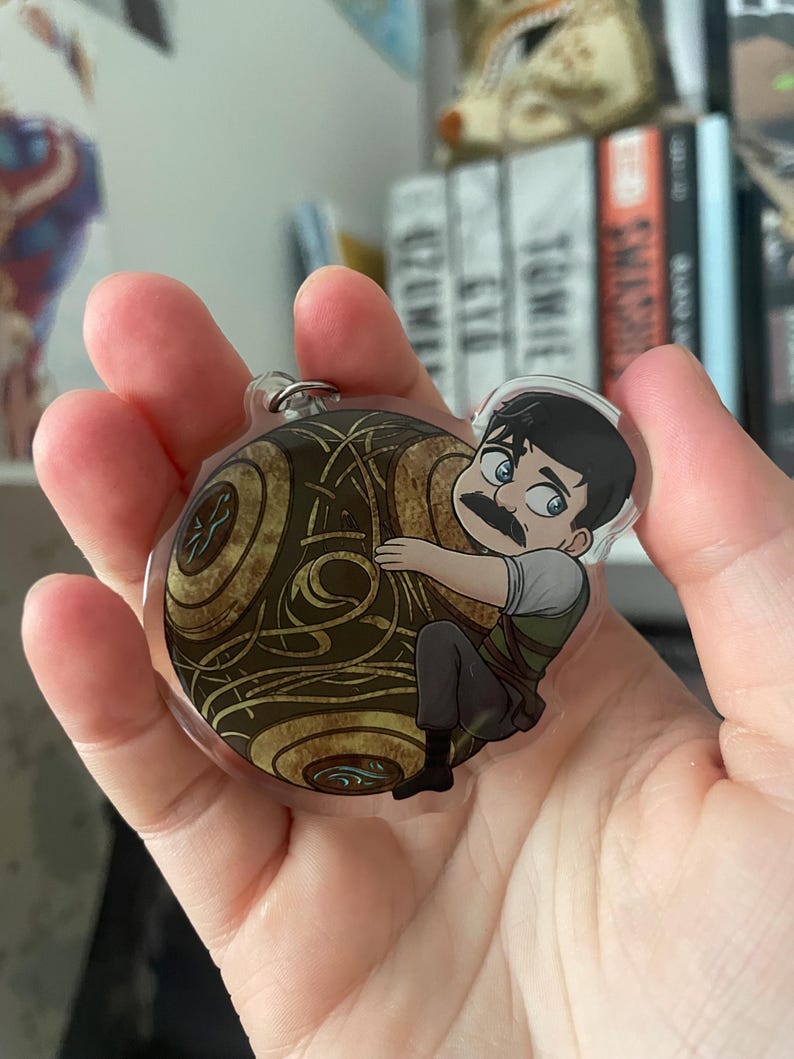 May include: A clear acrylic keychain featuring a cartoon illustration of a character with a mustache hugging a golden, patterned sphere. The keychain has a silver metal ring for attachment.