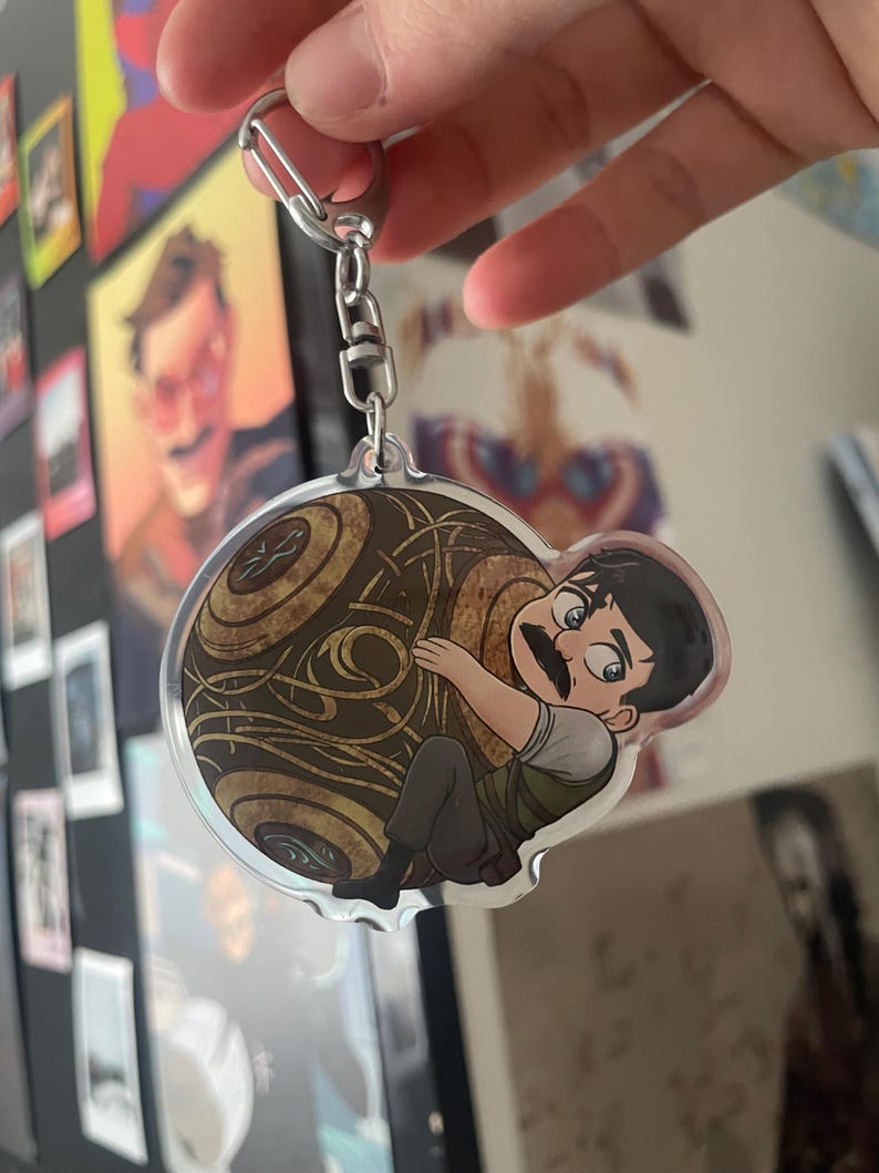 May include: A clear acrylic keychain featuring a cartoon character with a mustache hugging a brown, patterned sphere. The keychain has a silver metal clasp and chain. The character is wearing a green vest, gray shirt, and gray pants.