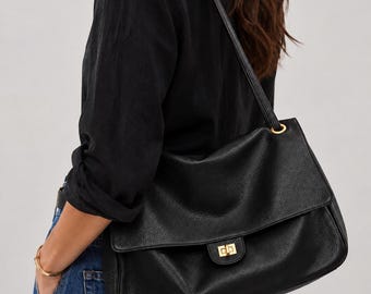 Puede incluir: Un bolso de hombro de cuero negro con textura granulada, que presenta una solapa, un cierre giratorio rectangular de tono dorado y una correa a juego sujeta con anillas de metal dorado en los laterales del bolso.