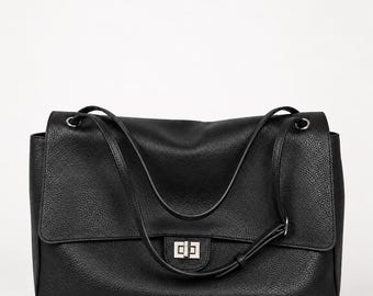 Puede incluir: Un bolso de hombro negro con una superficie texturizada y granulada. El bolso presenta un cierre de solapa rectangular asegurado por un mecanismo de cierre giratorio de metal plateado. Incluye dos correas finas y ajustables con herrajes plateados.