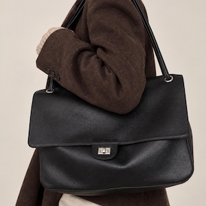 May include: A large black leather shoulder bag with a pebbled texture, featuring a rectangular flap, a silver-toned metal turn-lock closure, and two thin shoulder straps attached to the bag with silver-toned metal hardware rings.