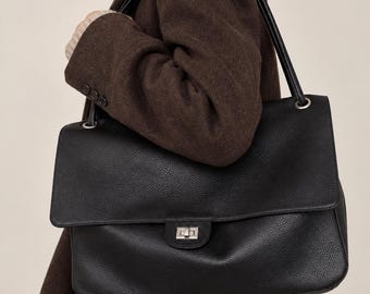 Puede incluir: Un bolso de hombro grande de cuero negro con textura granulada, que presenta una solapa rectangular, un cierre giratorio de metal plateado y dos correas finas para el hombro unidas al bolso con anillas de metal plateado.