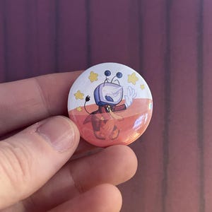 May include: A round button pin featuring a cartoon character with a TV head, wearing a red scarf and orange pants. The pin has a white background with yellow stars and a gradient of orange and pink.