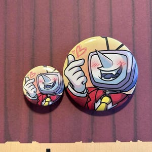 Puede incluir: Dos botones redondos con un personaje de dibujos animados. El personaje tiene una cabeza de televisión, un cuerno blanco y lleva una chaqueta roja y una corbata amarilla. El personaje está haciendo un corazón con los dedos. Un botón es más pequeño que el otro.