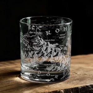 May include: A clear, cylindrical glass with a detailed white map design etched around its exterior. The map features mountain ranges and text, including "ROHA" and "ERED". The glass sits on a wooden surface.