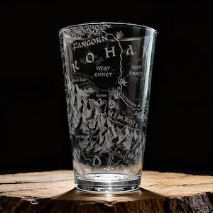 May include: A clear pint glass etched with a map of Rohan, including Fangorn, West Emnet, and Nimrais. The glass is cylindrical and sits on a wooden surface, with a dark background.