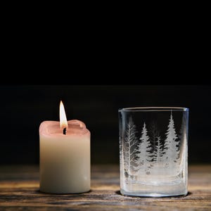 Può includere: Una candela a colonna bianca con una fiamma accesa accanto a un bicchiere di vetro trasparente con un disegno inciso di una fitta foresta con alti pini. Il bicchiere è cilindrico con un bordo liscio e una base spessa, con sagome di alberi.
