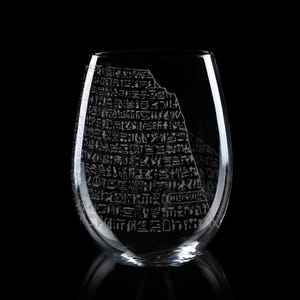 May include: A clear, stemless glass tumbler featuring a detailed, etched design of ancient Egyptian hieroglyphics arranged in a block-like pattern. The glass has a smooth, curved silhouette and a transparent finish, with the white-toned engravings standing out against the dark background.