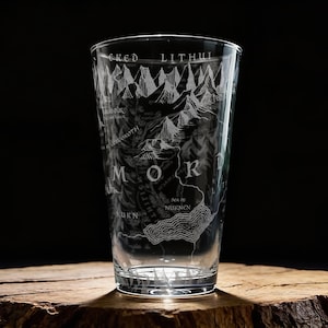 May include: A clear pint glass etched with a map of Mordor, featuring mountain ranges, text, and geographical details. The glass is cylindrical with a slightly flared top, perfect for enjoying beverages. The glass sits on a wooden surface.