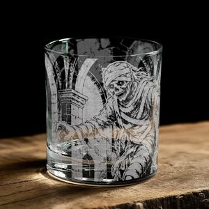 May include: A clear glass tumbler featuring a detailed white etched design of a skeletal mummy wrapped in bandages, positioned in front of stone architectural columns. The glass has a cylindrical shape with a thick base and smooth rim.