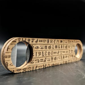 Peut inclure: Un décapsuleur en bois brun clair présentant une forme rectangulaire plate et allongée aux extrémités arrondies. La surface est gravée au laser avec des rangées d'hiéroglyphes égyptiens détaillés. Une découpe circulaire contient un mécanisme en métal.