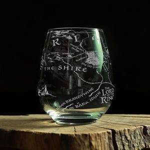 May include: A clear stemless wine glass featuring a white etched map of The Shire from The Lord of the Rings. The design includes detailed geographical labels such as Hobbiton, Buckland, The Old Forest, and River Brandywine, all rendered in a crisp, legible font.