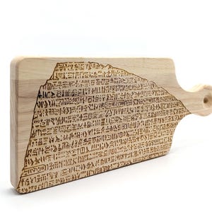 May include: A rectangular light-colored wooden cutting board featuring a laser-engraved design of ancient Egyptian hieroglyphics covering the surface. The board includes a handle on the right side with a circular hole for hanging or storage.