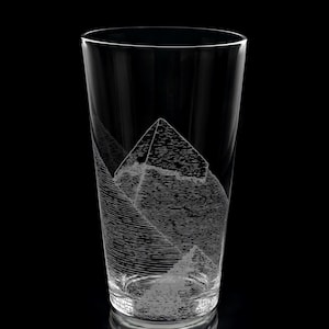 Peut inclure: Un verre transparent avec un motif gravé détaillé d'une pyramide et d'une chaîne de montagnes. Le motif est blanc sur le verre transparent. Le verre est cylindrique avec un dessus légèrement évasé. Le fond est noir.