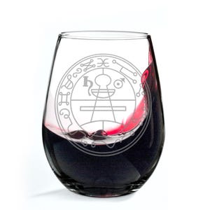 May include: A stemless wine glass with a detailed etched design. The glass is filled with red wine, with a splash visible. The etched design is a circular pattern with symbols and text. The glass is clear.