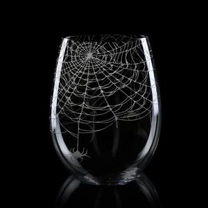 May include: A clear stemless wine glass featuring a white etched design of a detailed spider web covering the upper portion of the glass, with a small spider hanging from the web near the bottom edge of the etched pattern on the glass surface.