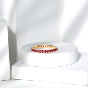 Può includere: Un anello a fascia in oro con una fila continua di piccole pietre preziose rotonde di un rosso vibrante, incastonate lungo l'intera circonferenza della fascia metallica. L'anello è esposto orizzontalmente su una piattaforma cilindrica bianca.