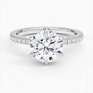 May include: A silver engagement ring with a large, round, clear diamond center stone. The band is also adorned with smaller, clear diamonds. The ring is set against a white background, highlighting its sparkle and design.