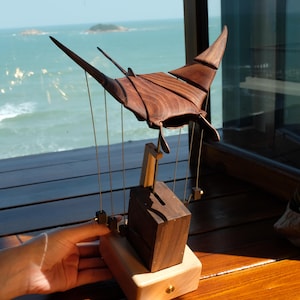 Puede incluir: Escultura de madera de una manta raya suspendida por cuerdas, con acabado marrón oscuro. Montada sobre una base de madera clara con un bloque rectangular marrón oscuro. El fondo muestra un paisaje marino.