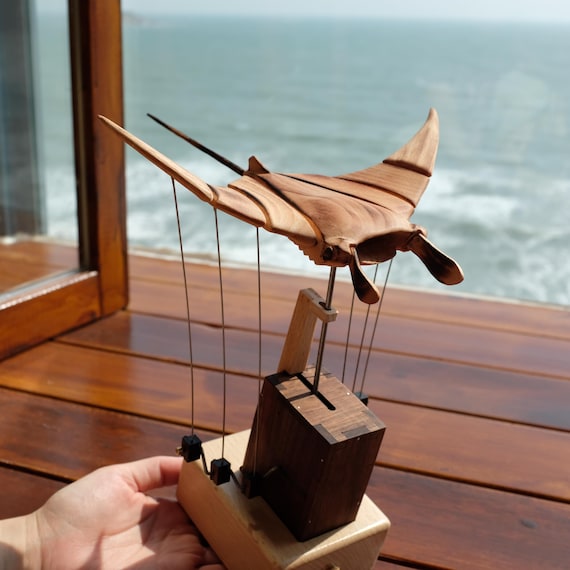 A hand-carved solid wood manta ray that actually flies/glides via a crank mechanism — kinetic sculpture meets ocean art, and no two are alike.