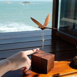 Puede incluir: Una escultura de pájaro de madera con las alas extendidas, suspendida sobre un pequeño cubo de madera oscura. El pájaro está sujeto al cubo con finos cables dorados. La escultura está situada frente a una ventana con vistas al océano.