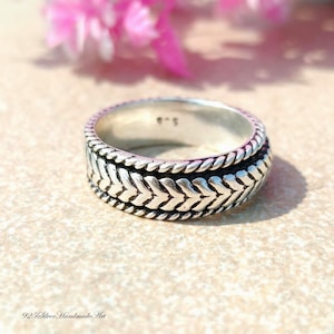 Könnte beinhalten: Ein silberner Ring mit einem strukturierten Fischgrätmuster. Der Ring hat ein Banddesign mit einem sich wiederholenden V-förmigen Motiv. Der Ring liegt auf einer neutralen Oberfläche mit rosa Blumen im Hintergrund. Der Text "925 Silver Handmade Art" steht unten.