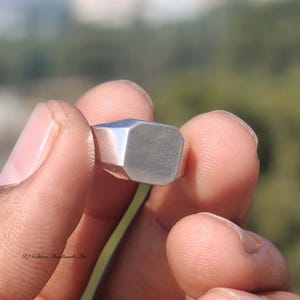 May include: A silver ring with a geometric, octagonal face. The ring is held between fingers, showcasing its polished surface. The ring's design is simple and modern, suitable for everyday wear. The ring is a classic piece of jewelry.